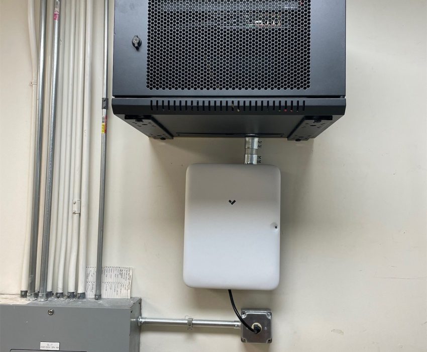 Verkada Installers A black wall-mounted server cabinet, suited for Verkada Licenses security system Verkada installers, is positioned above a white electrical box, with various pipes, cables, and a gray electrical panel visible on a beige wall. Verkada Installers A black wall-mounted server cabinet, suited for Verkada Licenses security system Verkada installers, is positioned above a white electrical box, with various pipes, cables, and a gray electrical panel visible on a beige wall.