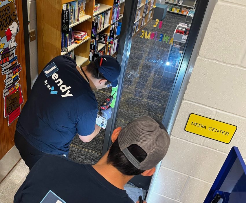 Verkada Installers Two people in Jendy shirts, likely Verkada installers, work on equipment outside a school media center near a mirror and bookshelves. One kneels with tools while the other bends over an open blue case with a water bottle inside. Verkada Installers Two people in Jendy shirts, likely Verkada installers, work on equipment outside a school media center near a mirror and bookshelves. One kneels with tools while the other bends over an open blue case with a water bottle inside.